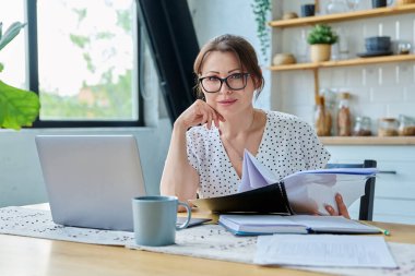 Orta yaşlı gülümseyen güzel bir kadın dizüstü bilgisayar belgeleriyle çalışıyor, mutfaktaki masada oturan kameraya bakıyor. Teknoloji, iş, iş, olgun insanlar konsepti