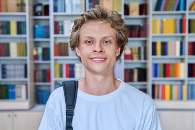 Kütüphanede kameraya bakan yakışıklı, gülümseyen bir gencin portresi. 16-17 yaşlarında, sırt çantalı, kitap raflarıyla sınıfta poz veren genç bir erkek. Eğitim ergenlik bilgisi