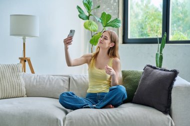 Gülümseyen genç bayan akıllı telefonuyla selfie çekiyor, evdeki kanepede oturuyor. Eğlence, eğlence, gençlik, yaşam tarzı konsepti