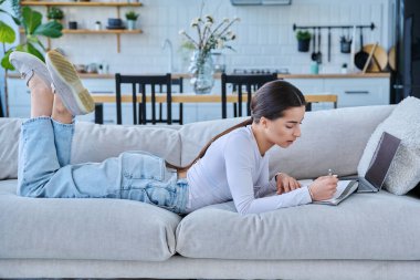 Evde dizüstü bilgisayarla kanepede uzanmış sınav için notlar alan genç üniversiteli kız. Eğitim e-öğrenim kursu online ders yaşam tarzı gençlik kavramı