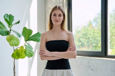 18-20 yaşlarında gülümseyen genç bir kadının portresi pencerenin yanında çapraz kollarla kameraya bakıyor. Uzun düz saçlı, zarif sarışın kız. Gençlik yaşam tarzı güzellik modası