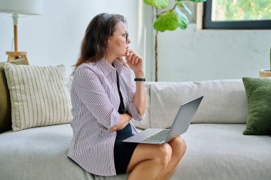 Mutsuz, ciddi, orta yaşlı bir kadının koltukta dizüstü bilgisayar kullanması üzücü. Kötü haberler, iş hayatında sorunlar, vücut ağrısı, akıl sağlığı konsepti