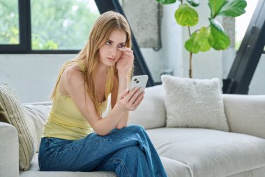 Üzgün, ağlayan, akıllı telefona bakan, evdeki kanepede oturan, fotokopi çeken genç bir kadın. İletişim kurmak için telefon kullanan bir kadın, eğitim için mobil uygulamalar sosyal ağlar
