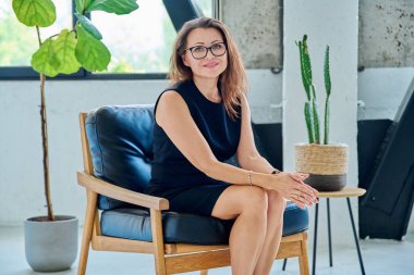 Sandalyede oturan, rahat gülümseyen ve kameraya bakan zarif, başarılı orta yaşlı iş kadınının portresi. İş, iş, yaşam tarzı, olgun insanlar konsepti