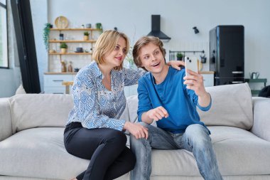 Orta yaşlı anne ve genç oğul evde oturup akıllı telefondan selfie çekiyorlar. Yaşam tarzı, ebeveyn-ergen ilişkisi, aile sağlığı, ergen yaşı kavramı.