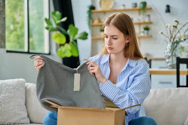Karton kutuda paketleri açan üzgün genç bir kadın sipariş edilmiş kıyafetlere bakıyor, kanepede oturuyor. E-alışveriş, e-market, teslimat servisi, online kıyafet siparişi.