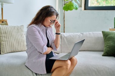 Mutsuz, ciddi, orta yaşlı bir kadının koltukta dizüstü bilgisayar kullanması üzücü. Kötü haberler, iş hayatında sorunlar, vücut ağrısı, akıl sağlığı konsepti