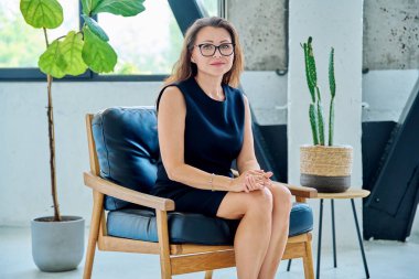 Sandalyede oturan, rahat gülümseyen ve kameraya bakan zarif, başarılı orta yaşlı iş kadınının portresi. İş, iş, yaşam tarzı, olgun insanlar konsepti