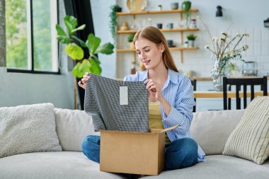 Karton kutuda paketleri açan mutlu genç bir kadın sipariş edilmiş kıyafetlere bakıyor, kanepede oturuyor. E-alışveriş, e-market, teslimat servisi, online kıyafet siparişi.