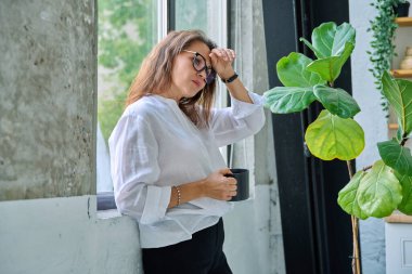 Olgun, üzgün, bunalımlı, stresli bir kadın, pencerenin yanında duran zihinsel ve fiziksel acı. Sorunlar yalnızlık, baş ağrısı hormon stresi, orta yaşlı kadınların sağlığı