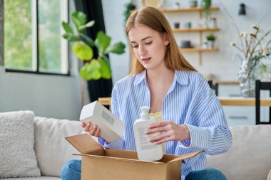 Genç, mutlu, karton kutuları internet siparişiyle açan, cilt bakımı parfümlü ev yapımı kimyasallar alan, evinde kanepede oturan bir kadın. E-alışveriş, e-market, teslimat servisi.