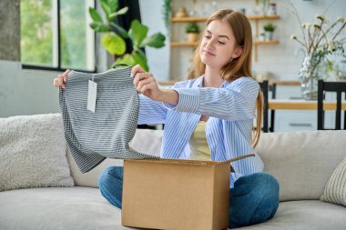 Karton kutuda paketleri açan üzgün genç bir kadın sipariş edilmiş kıyafetlere bakıyor, kanepede oturuyor. E-alışveriş, e-market, teslimat servisi, online kıyafet siparişi.