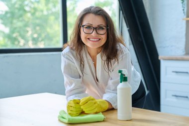 Gülümseyen orta yaşlı, eldivenli, bez temizleme spreyli güzel bir kadın kameraya bakıyor. Temizlik, temizlik, insanlar, yaşam tarzı konsepti