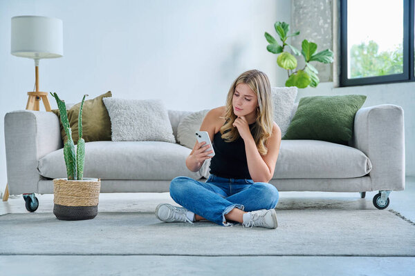 Улыбаясь довольной счастливой молодой женщине со смартфоном, сидящей дома рядом с диваном, женщина читает по мобильному телефону. Мобильные цифровые приложения для досуговой коммуникации