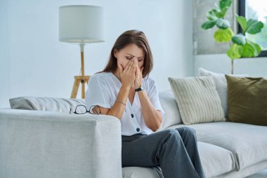 Olgun, üzgün, stresli bir kadın. Stresli, zihinsel ve fiziksel bir acı yaşıyor. Kanepede oturuyor. Sorunlar yalnızlık, baş ağrısı hormon stresi, orta yaşlı kadınların sağlığı