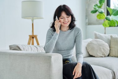 Güzel, pozitif, rahat, genç bir kadın cep telefonuyla konuşuyor, evdeki oturma odasında kanepede oturuyor. Konuşma, iletişim, cep telefonu, yaşam tarzı, gençlik kavramı