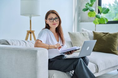 Orta yaşlı güzel bir kadının portresi, dizüstü bilgisayarlı koltukta oturuyor, rahatlamış gülümseyen bir kadın kameraya bakıyor. Teknoloji, yaşam tarzı, iş, eğlence, insan kavramı