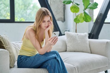 Üzgün, üzgün, genç bir kadın akıllı telefona bakıyor. Evdeki koltukta oturuyor, fotokopi çekiyor. İletişim kurmak için telefon kullanan bir kadın, eğitim için mobil uygulamalar sosyal ağlar
