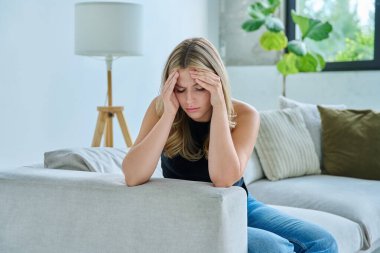 Mutsuz, üzgün, başı ağrıyan genç bir kadın, evdeki kanepede oturuyor. Duygusal fiziksel acı çeken üzgün bir kadın. Sorunlar ruhun ve bedenin sağlığı ve gençliğin sorunları
