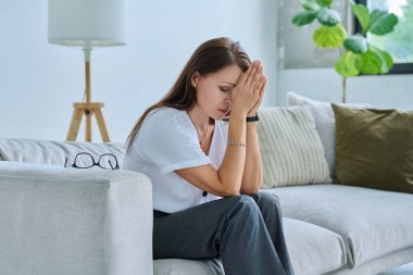 Olgun, üzgün, stresli bir kadın. Stresli, zihinsel ve fiziksel bir acı yaşıyor. Kanepede oturuyor. Sorunlar yalnızlık, baş ağrısı hormon stresi, orta yaşlı kadınların sağlığı