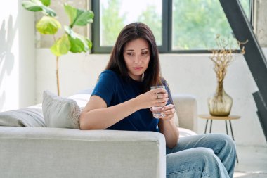 Evdeki kanepede oturan, elinde cam suyla üzgün genç bir kadın. Depresyon, stres, acı, sorunlar, akıl sağlığı insanları kavramı