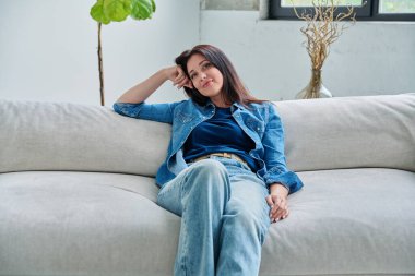 Oturma odasındaki kanepede oturan güzel, mutlu, esmer kadının portresi. 30 'lu yaşlarda çekici bir kadın kameraya bakıyor. Güzellik yaşam tarzı rahatla rahat rahat mutluluk, kadın konsepti