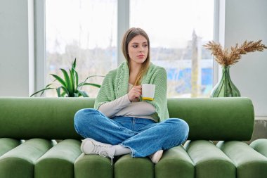 Evdeki kanepede elinde fincanla oturan rahat genç bir kadının portresi. Çekici bir kadın oturma odasında kupayla kanepede dinleniyor. Boş zaman mutluluğu rahat yaşam tarzı gençlik konsepti