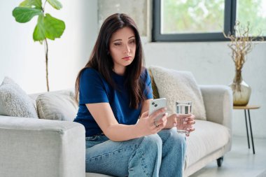 Evdeki kanepede oturan üzgün ve mutsuz genç bir kadın akıllı telefon kullanıyor. İletişim, eğlence, alışveriş, iş ve eğitim için mobil başvurular. Teknoloji yaşam tarzı duyguları insanların konsepti