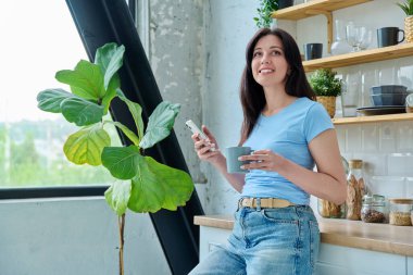 Güzel, mutlu, gülümseyen, 30 'lu yaşlarda, evinde kameraya bakarak akıllı telefon kullanan bir kadının portresi. Yaşam tarzı, ev hayatı, insan kavramı.