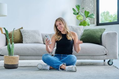Video sohbet konferansı için akıllı telefonu kullanan gülümseyen genç bir kadın evde oturmuş web kamerasına, telefon ekranına bakıyor. Eğitim teknolojileri eğlence iletişimi, gençlik kavramı