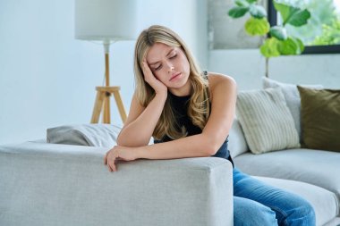 Mutsuz, üzgün, başı ağrıyan genç bir kadın, evdeki kanepede oturuyor. Duygusal fiziksel acı çeken üzgün bir kadın. Sorunlar ruhun ve bedenin sağlığı ve gençliğin sorunları