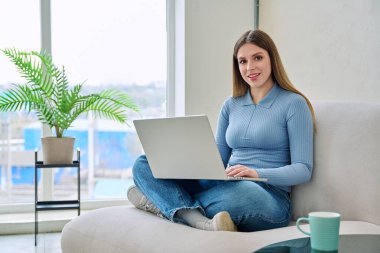 Evde dizüstü bilgisayarla oturmuş kameraya bakan genç bir kadının portresi. Güzel bir kadın iletişim eğitimi için bilgisayar kullanıyor alışveriş eğitimi iş teknolojisi yaşam tarzı