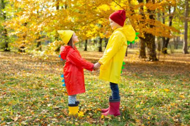 Sonbahar parkında su geçirmez yağmurluk ve lastik çizmeli çocuklar el ele tutuşur.