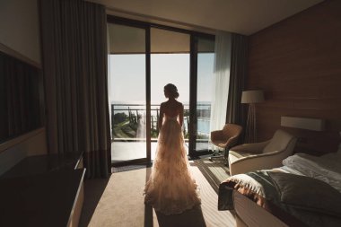 Gelin, otel odasındaki yumuşak ışıkta pencere balkonunu açıyor. Beyaz elbiseli, muhteşem şehvetli gelinin arkasında