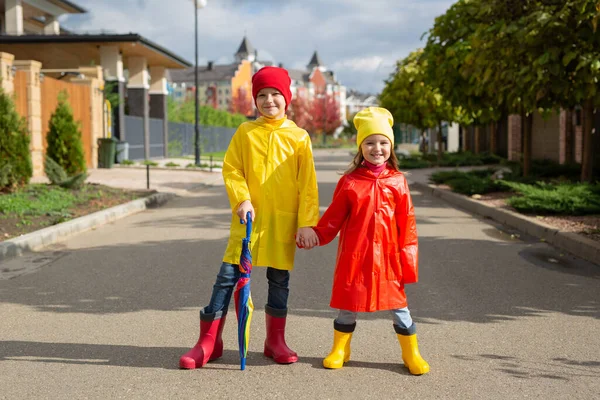 Stock image Two charming children, a boy and a girl, in waterproof raincoats and rubber boots with an umbrella