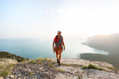 Elinde teçhizatla bir dağın tepesinde, denize bakan bir dağcı.