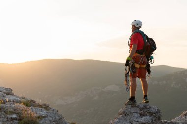 Elinde bir ip ve ekipmanla dağcı gün batımında dağın tepesinde duruyor.