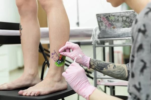 Tattoo artist puts a drawing of a ladybug on the leg of a young woman, the process of creating a tattoo. A girl takes a picture on a womans leg. A tattoo artist makes a tattoo. Close-up.