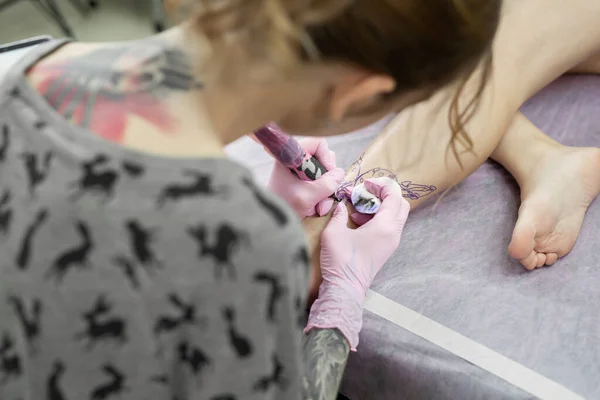 タトゥースタジオで入れ墨をするマスター プロのタトゥー師はスタジオで働いている ピンクの手袋の女性は若い女の子の足に入れ墨をします — ストック写真