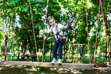 Tırmanma ekipmanlı küçük bir çocuk bir macera parkında halat ve karabinaya tutunarak ip boyunca yürüyor..