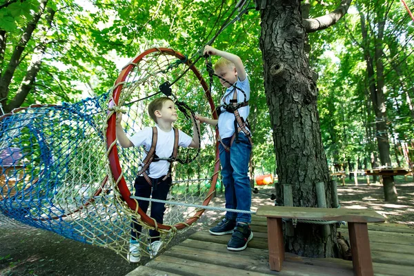 Two little boy in climbing gear is walking along a rope road in an adventure Park, holding onto a rope and a carabiner.