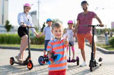 Mutlu bir aile parkta elektrikli scooter ve jiroskopla gezer. Ön planda küçük bir çocuk elinde bir paten tutar ve baş parmağını kaldırır.