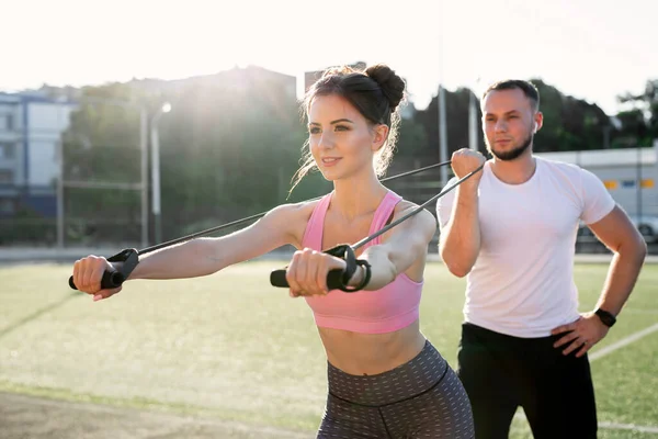 stock image Close-up of young sports people training at the stadium in the summer, a woman doing exercises with an expander and a man helping her