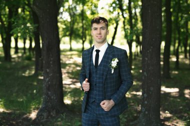 Doğada kameranın önünde poz veren mutlu bir damadın portresi. Arka planda ağaçlar var..