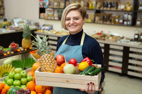 Lächelnde Verkäuferin Hält Eine Holzkiste Mit Gemüse Und Obst Laden lizenzfreie Stockfotos