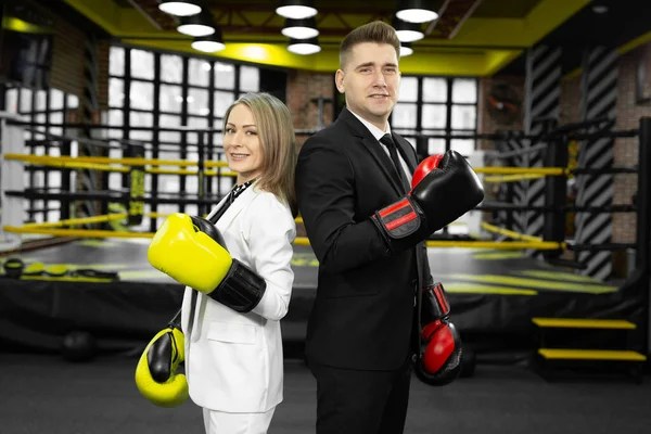 Man and woman in a suit and boxing gloves stand in front of the ring with their backs to each other