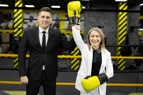 Colleagues, a man and a woman in business suits and boxing gloves in the ring. The man raises the womans hand as a winner.