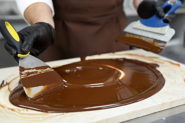 Male chocolatier uses a spatula to stir the tempered liquid chocolate on a granite table