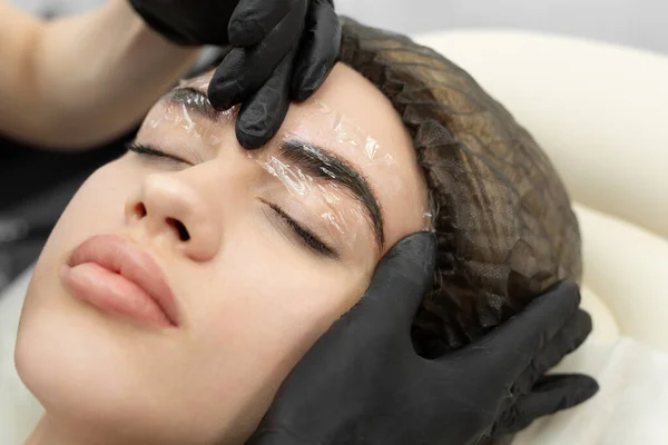 Girls face with closed eyes close-up. A white cream is applied on the eyebrows - an analgesic and the cosmetologists hands cover the eyebrows with a transparent film.