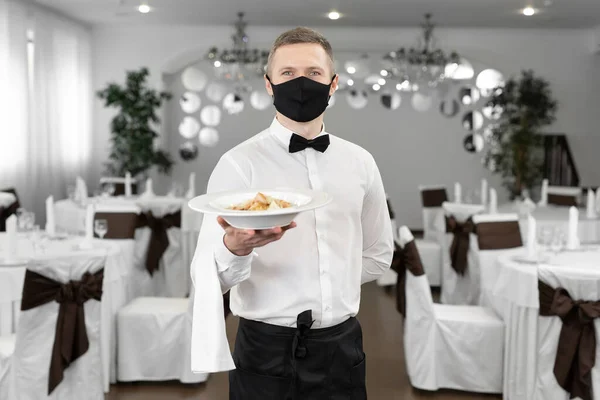 Young happy waiter wearing protective face mask while serving food in a restaurant